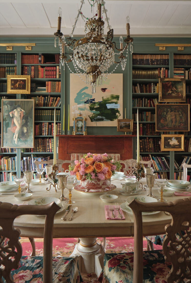 A dining room with dark sage green bookshelves, paiuntings, a crystal chandelier, and a round set table with a bouquet