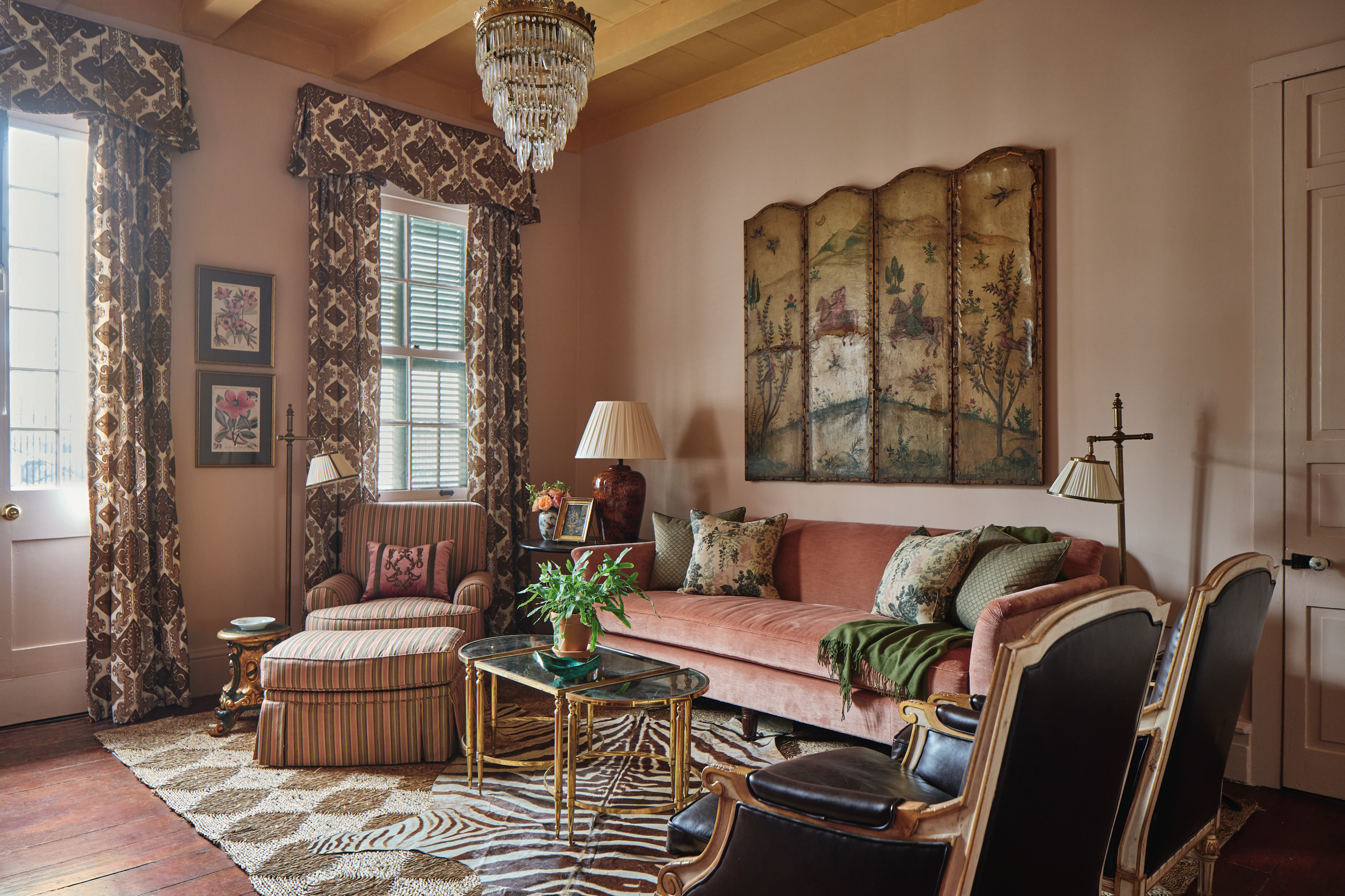 A pink living room with a tiered crystal chandelier, a painted leather screen, a velvet pink sofa, and chairs on a patterned diamond rug