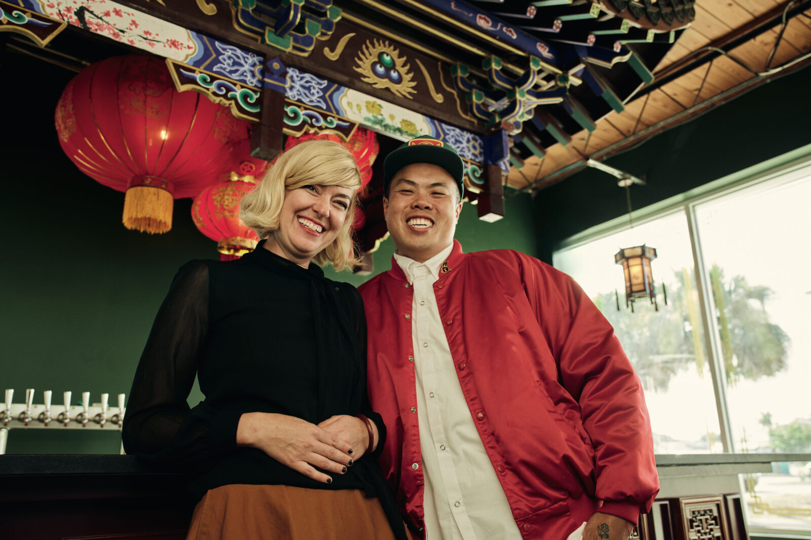 A woman in a black top and a man in a red jacket smile in front of a Chinese bar with ornate patterning and a green wall
