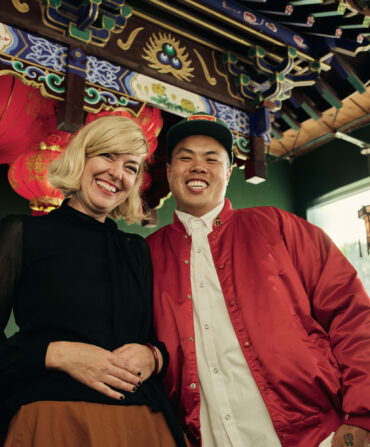 A woman in a black top and a man in a red jacket smile in front of a Chinese bar with ornate patterning and a green wall