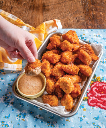 A hand dips a chicken nugget into light orange sauce. There are chicken nuggets on a silver platter on top of a bloral blue tablecloth