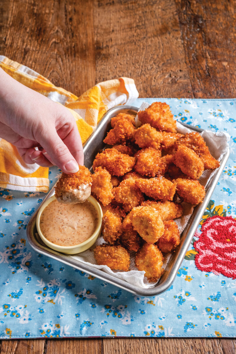 A hand dips a chicken nugget into light orange sauce. There are chicken nuggets on a silver platter on top of a bloral blue tablecloth