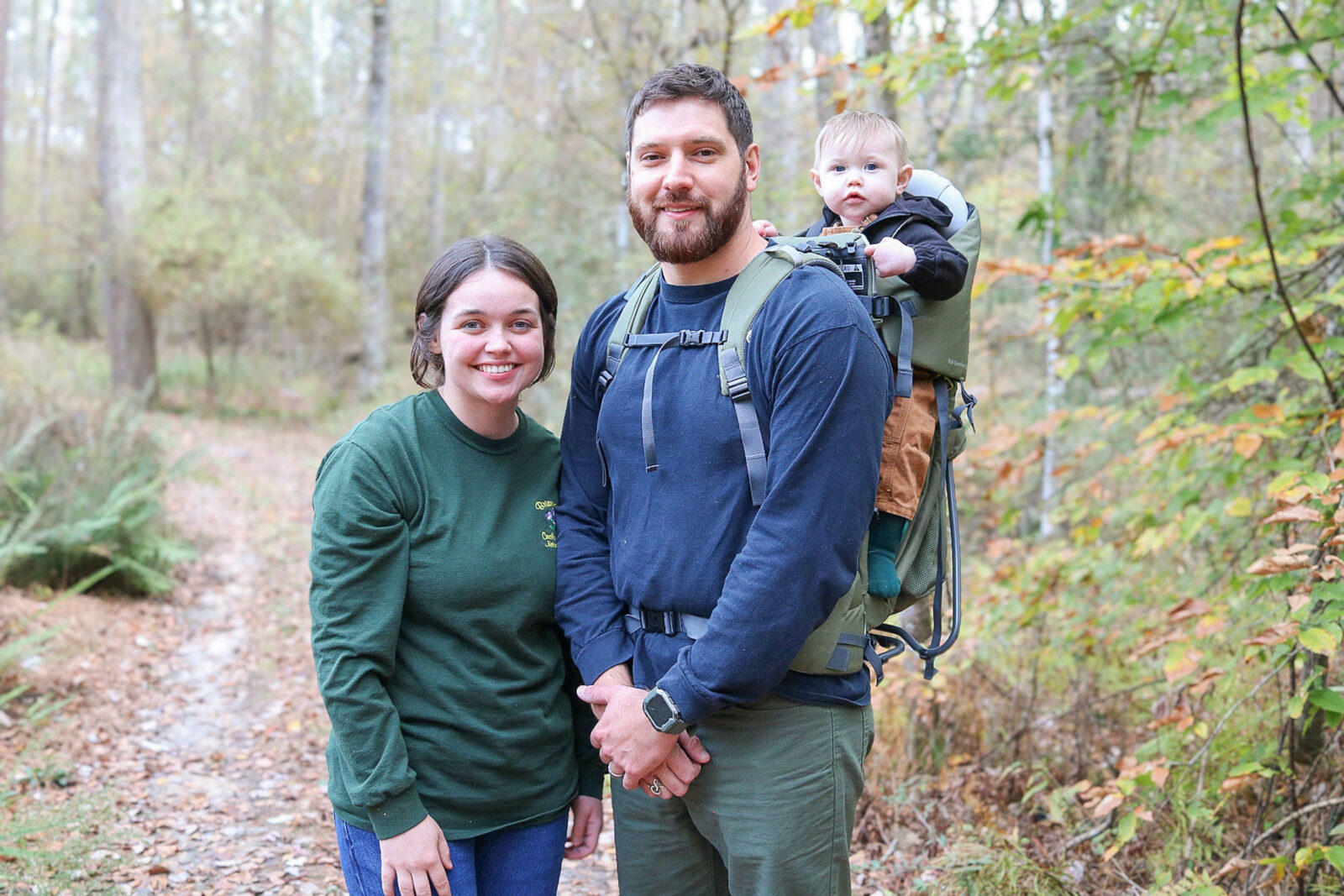 A woman and man with a baby stand in the woods
