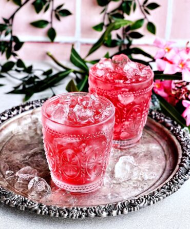 Two icy pink drinks in ornate glassware sit on a silver platter. There are flowers, vines, and pink tiles behind the drinks.