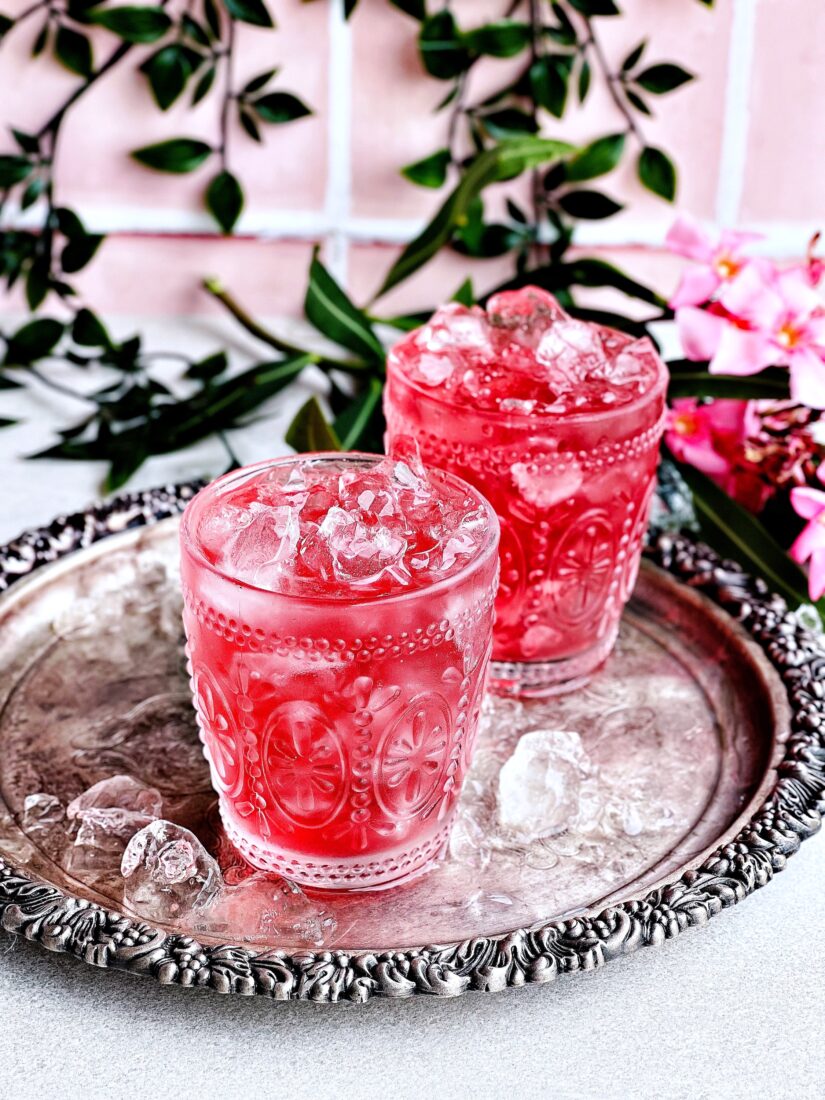 Two icy pink drinks in ornate glassware sit on a silver platter. There are flowers, vines, and pink tiles behind the drinks.