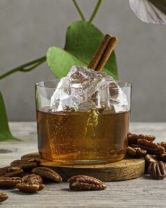 An old fashioned cocktail with a cinnamon stick sits on a wooden circle surrounded by walnuts