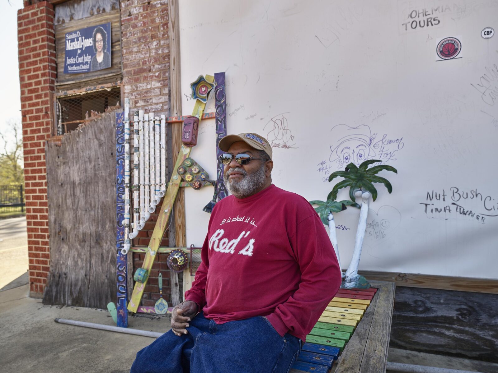 A man sitting down on a rainbow xylophone wearing a red sweatshirt that says "It is what it is... Red's" on it.