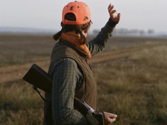 A girl with an orange hunting hat and camo raises a hand in the air; she has a gun under her arm