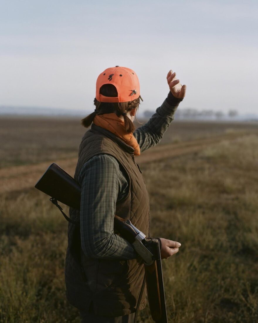 A girl with an orange hunting hat and camo raises a hand in the air; she has a gun under her arm