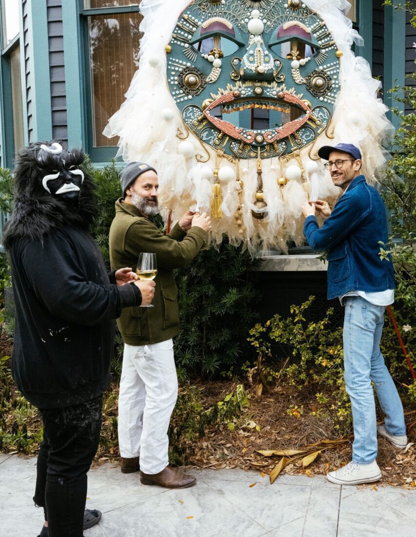 Two men set up a huge mask sculpture outside a house; a man in a black costume and face masks stands on the left