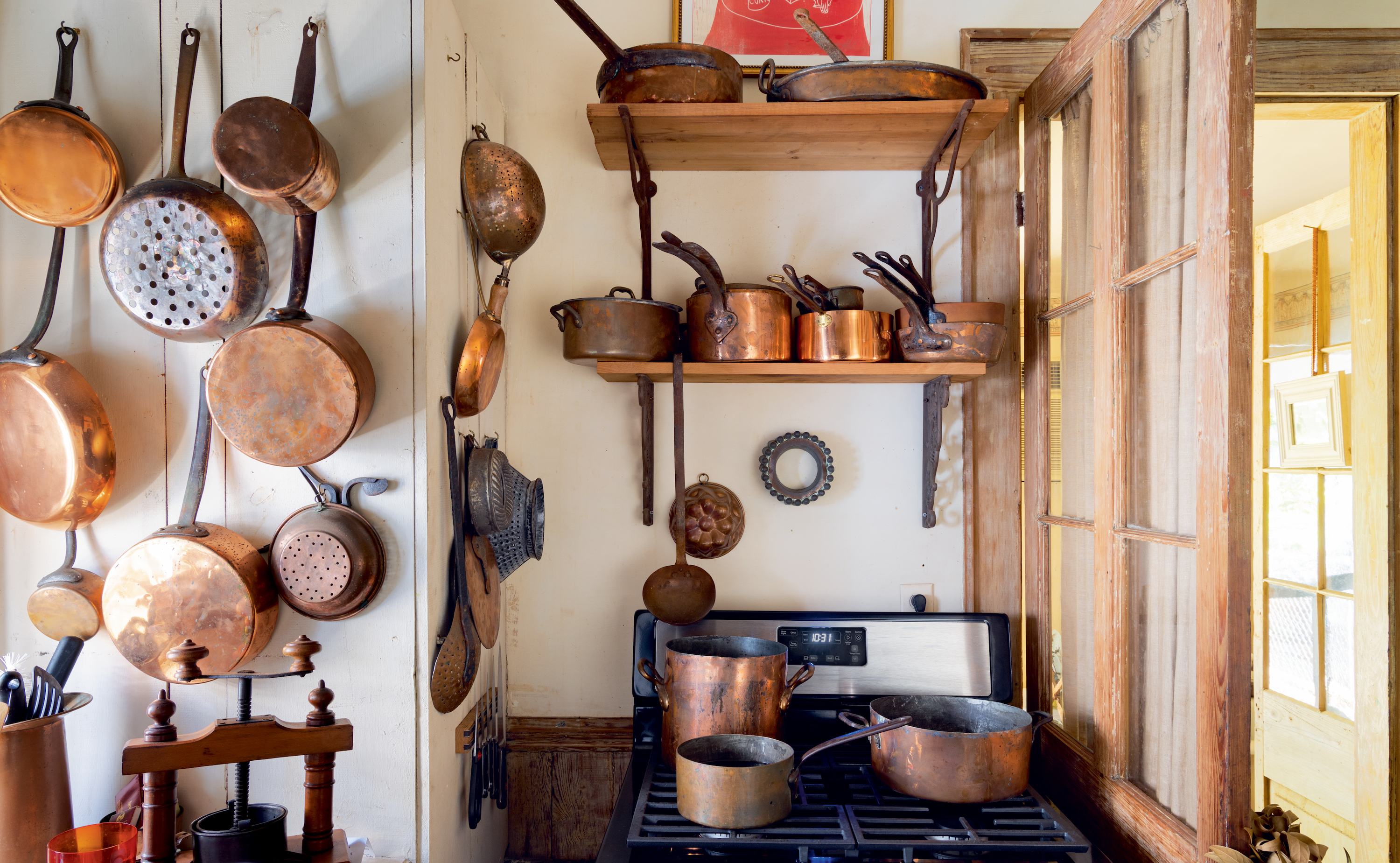 A kitchen with copper pots on the walls and on a stove. On the right, a wood door is open