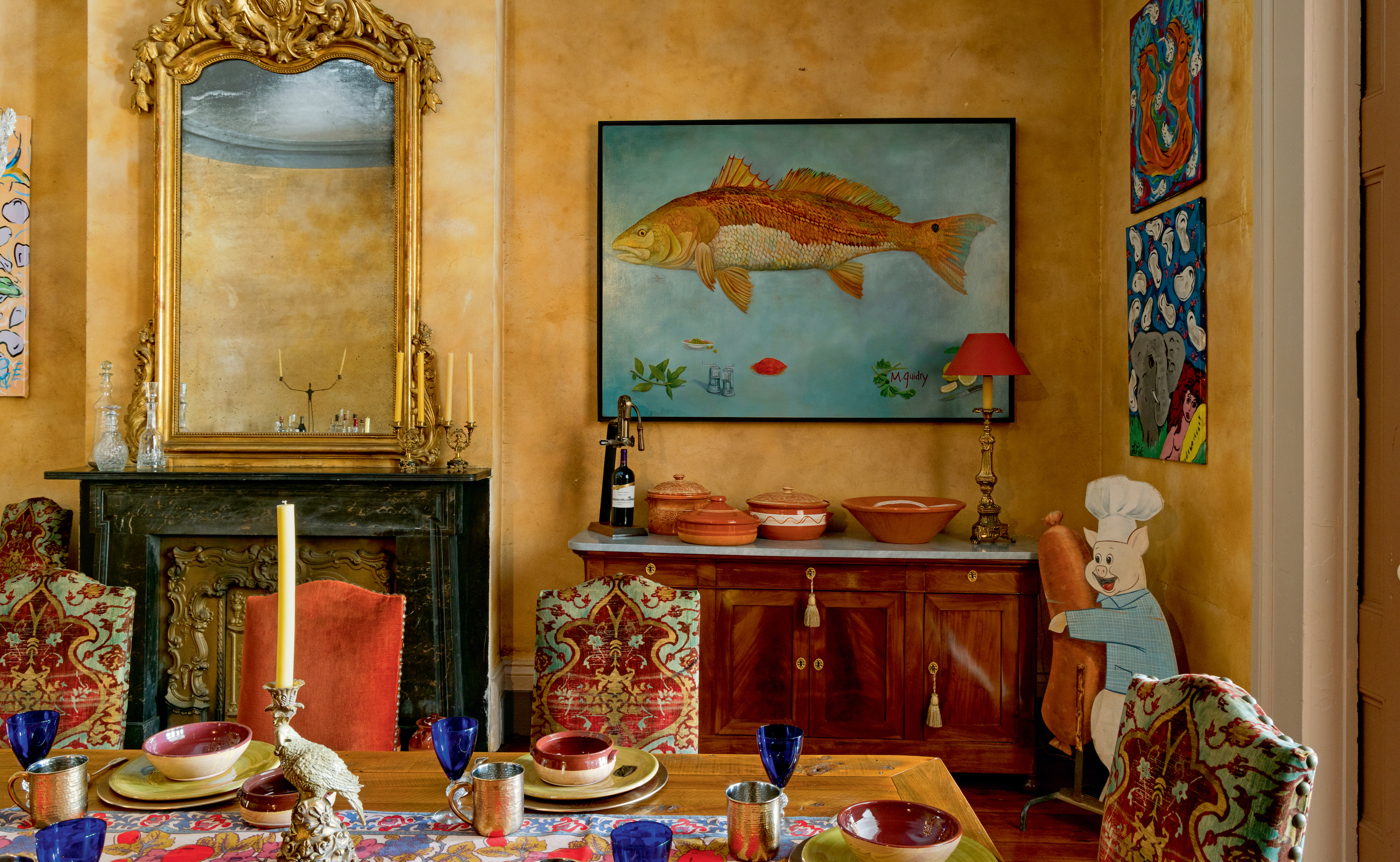 A yellow dining room with eclectic art, a wood dining table with patterned chairs, a fireplace, and a wood cabinet. A blue and brown painting of a fish hangs on the wall.