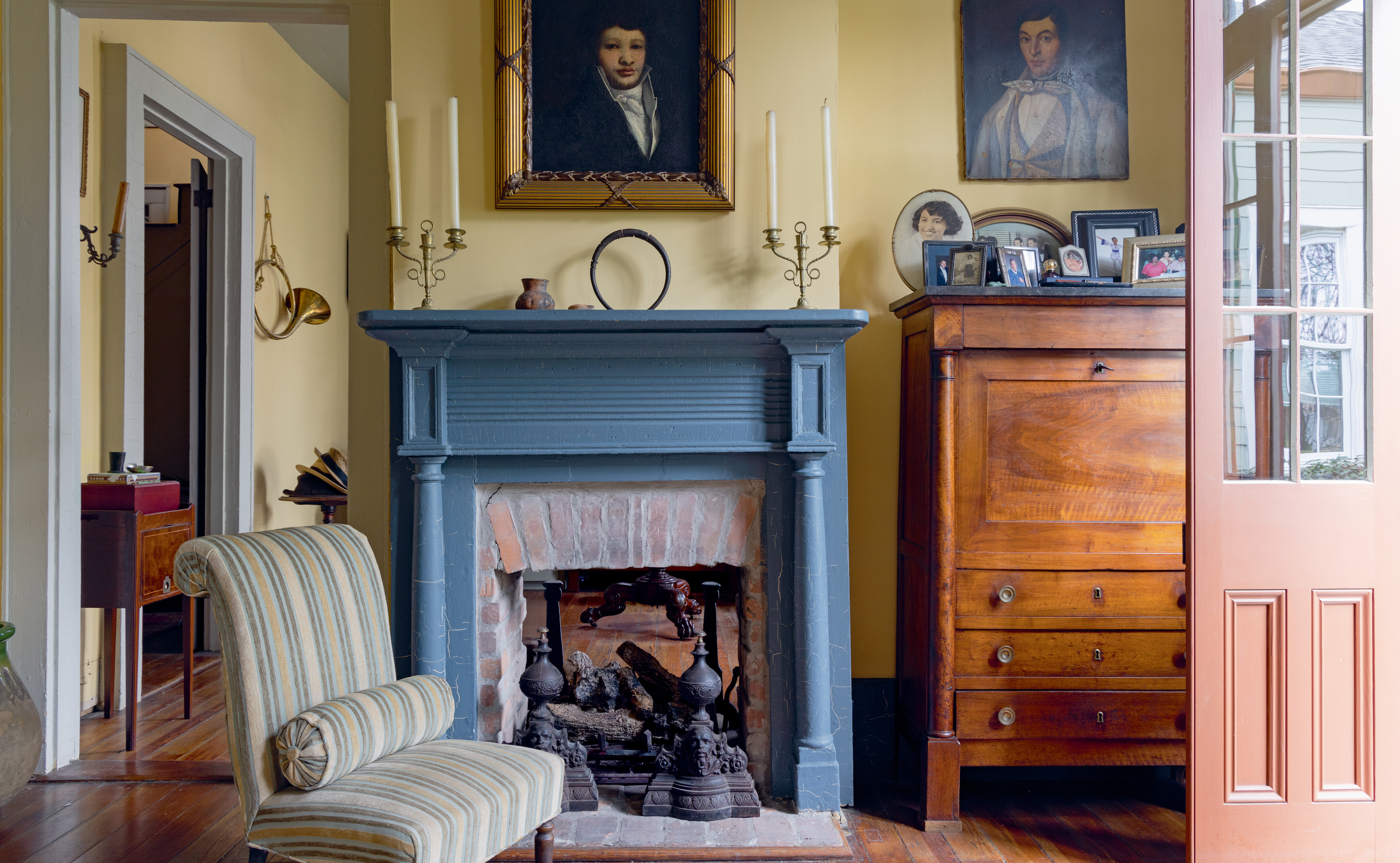 A blue mantle in a butter yellow living room. There are oil portraits of men and candle sticks decorating the room,