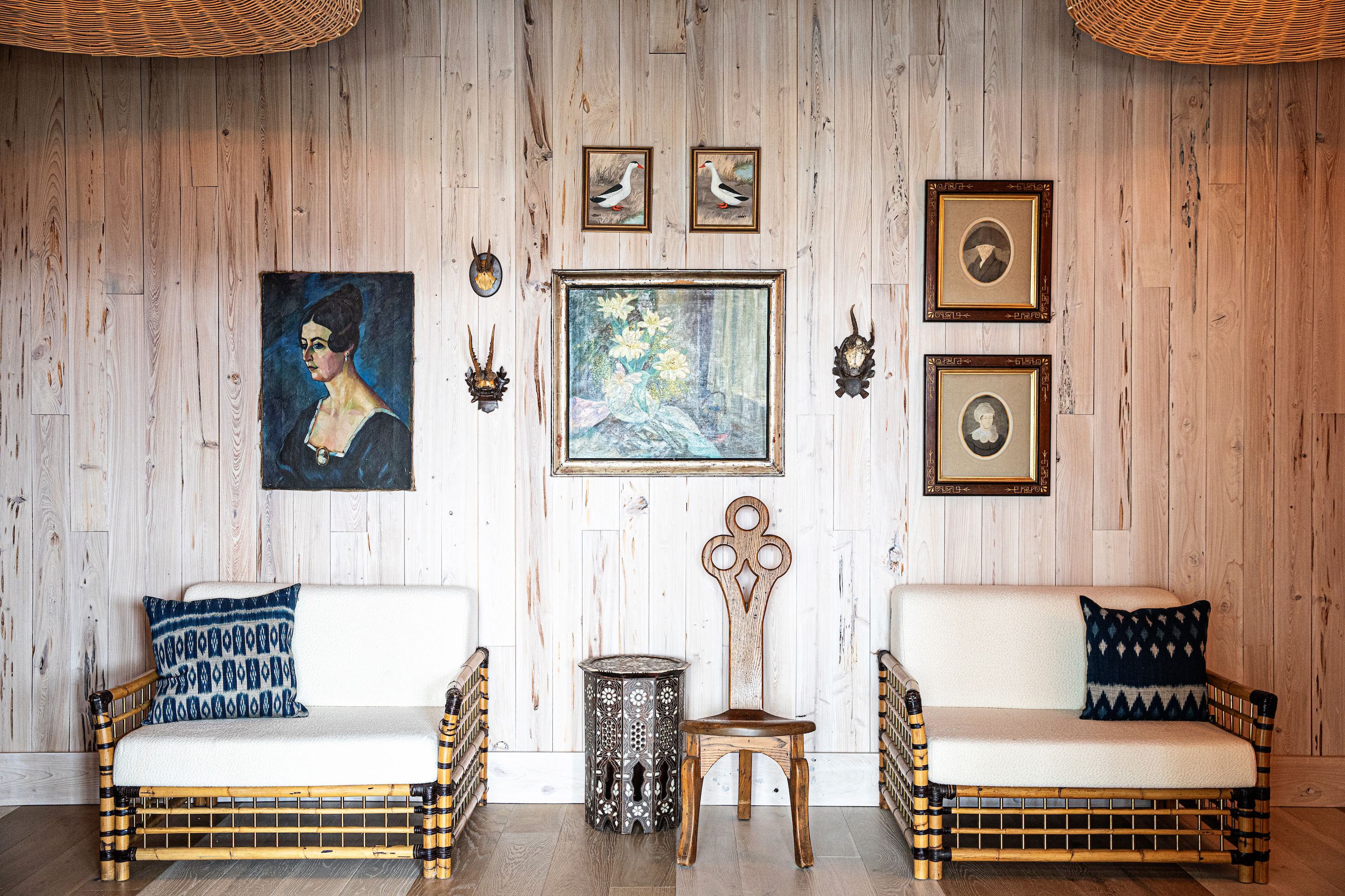 Antique art hangs on a wood wall above two wide chairs.
