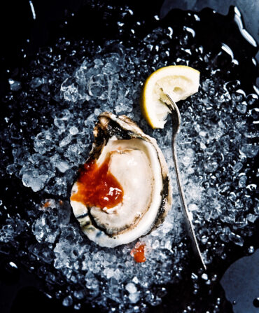 An oyster with cocktail sauce sits on a bed of crushed ice next to a lemon wedge with a fork in it.