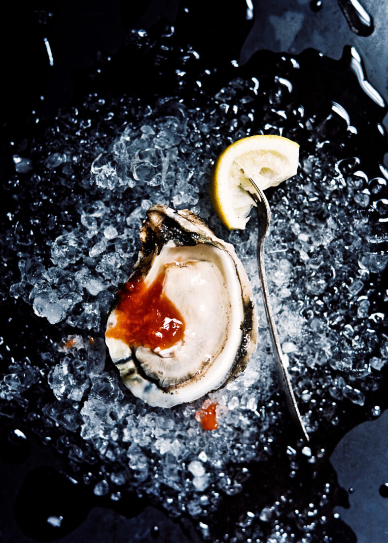 An oyster with cocktail sauce sits on a bed of crushed ice next to a lemon wedge with a fork in it.