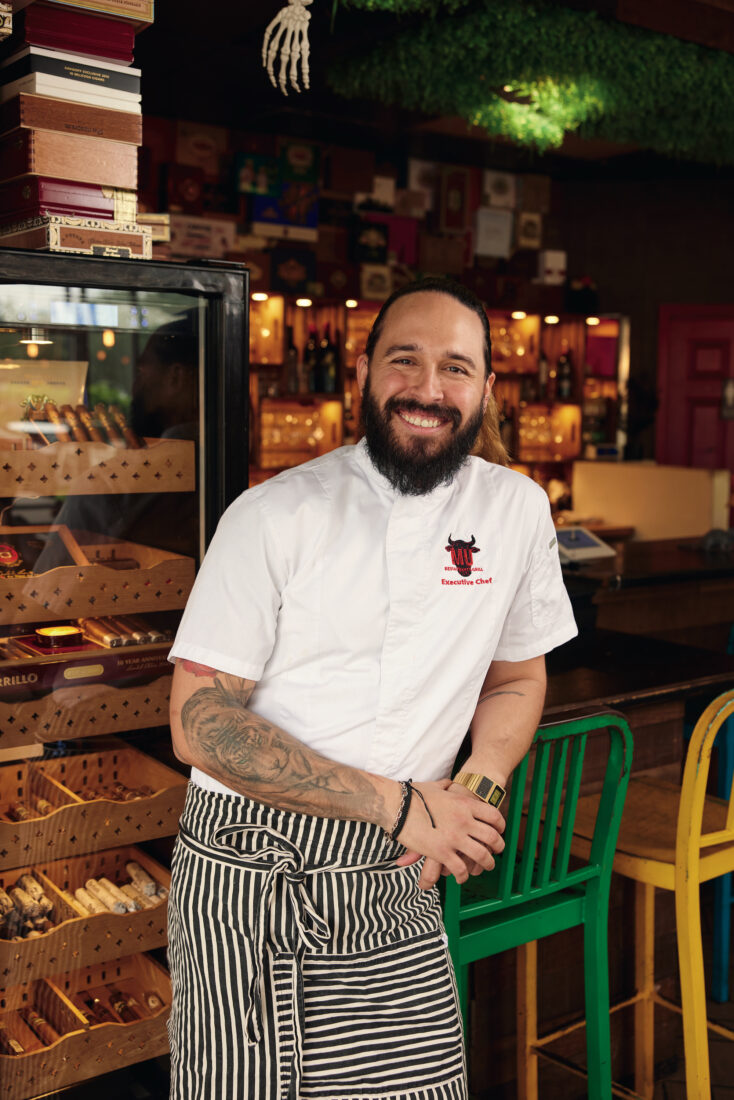 A portrait of a chef wearing a white shirt and striped apron. He stands against a bar and smiles