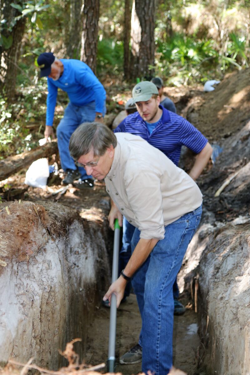 Three men study an excavated canal ditch in the dirt and stand in it