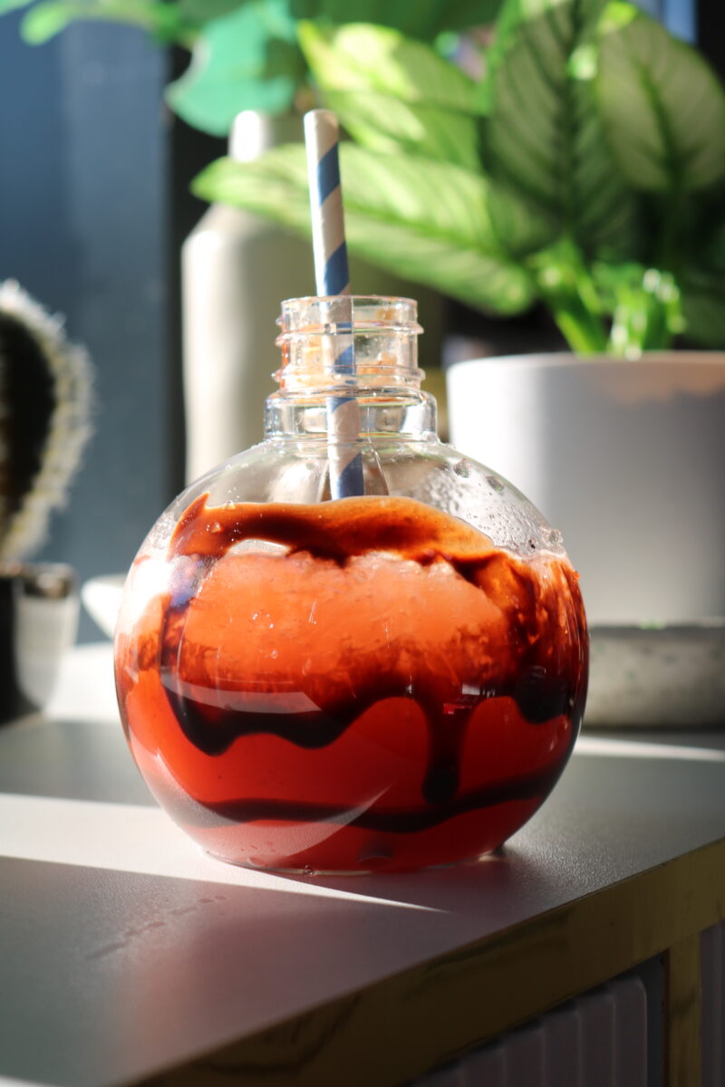 A circular glass cup with chocolate drizzle and strawberry juice inside. It has a striped blue and white straw