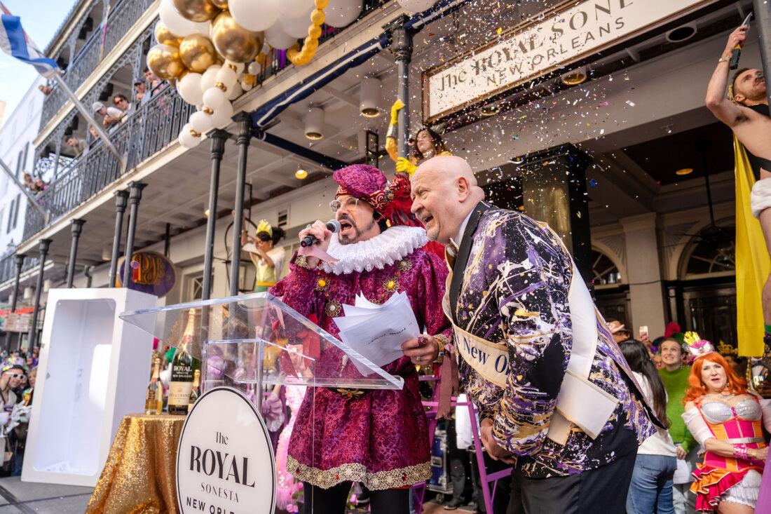 Two men dressed up for Mardi Gras stand in front of poles by a building with gold and white balloons