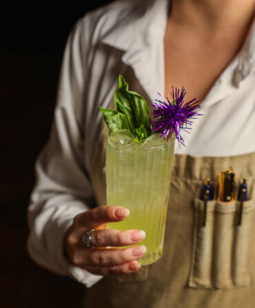 A hand holds a tall green cocktail in a glass garnished with a purple sparkler stick and basil leaves