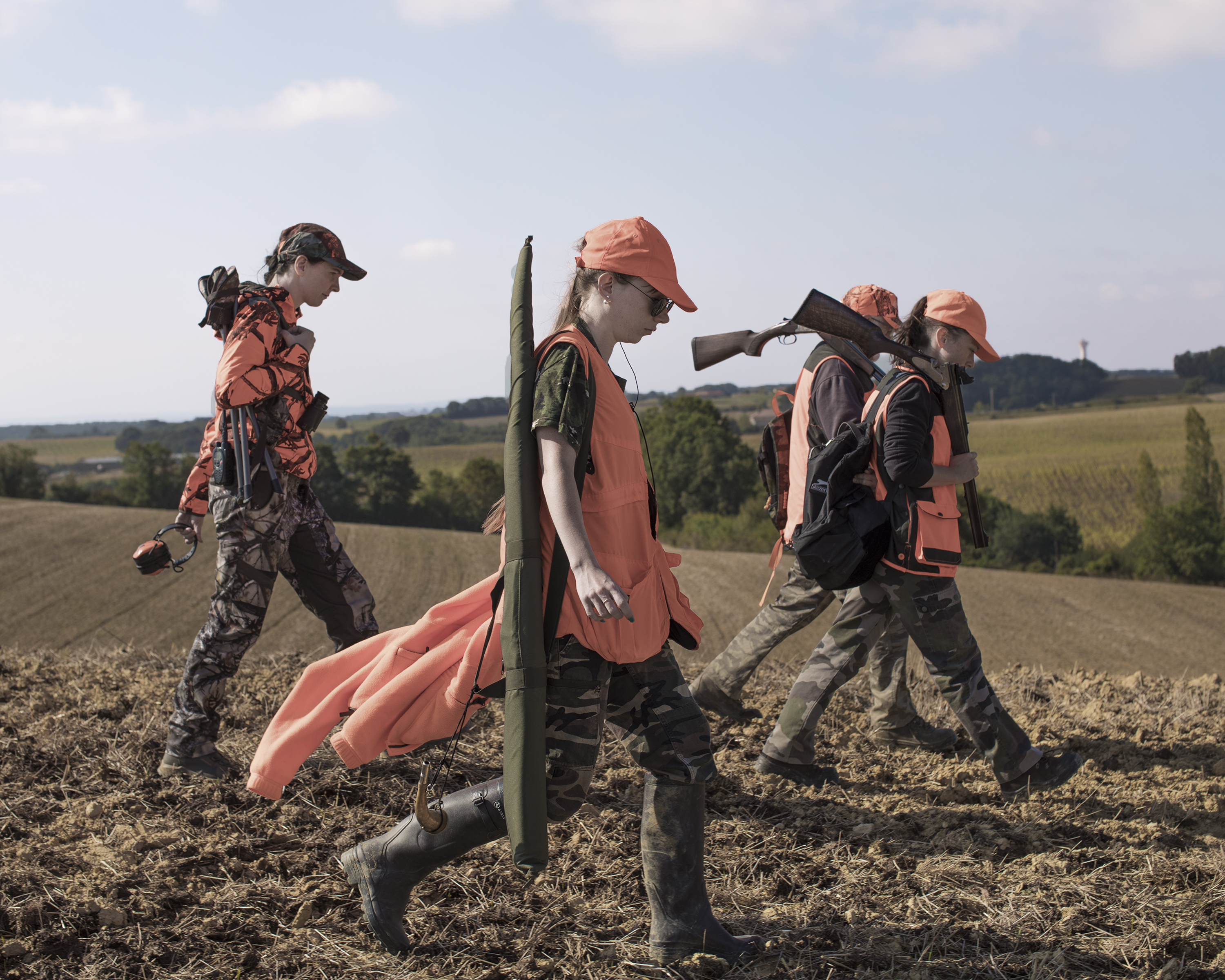 A group of women in orange hunting gear hold guns and walk through a field