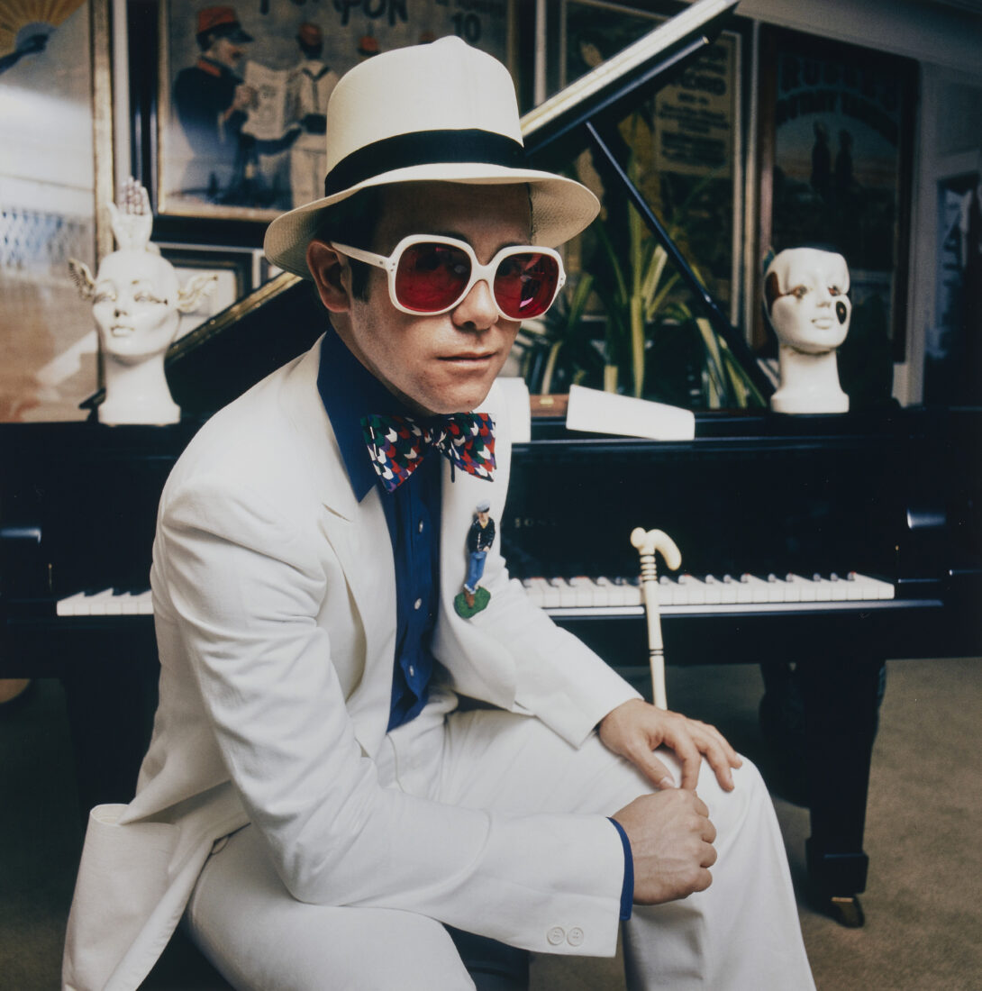 A photo of young Elton John in a white suit and hat holding a staff by a piano