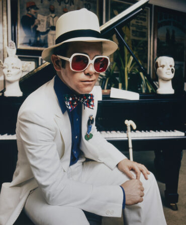 A photo of young Elton John in a white suit and hat holding a staff by a piano