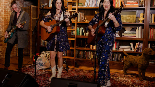 The Watson Twins playing their guitars in front of a bookshelf