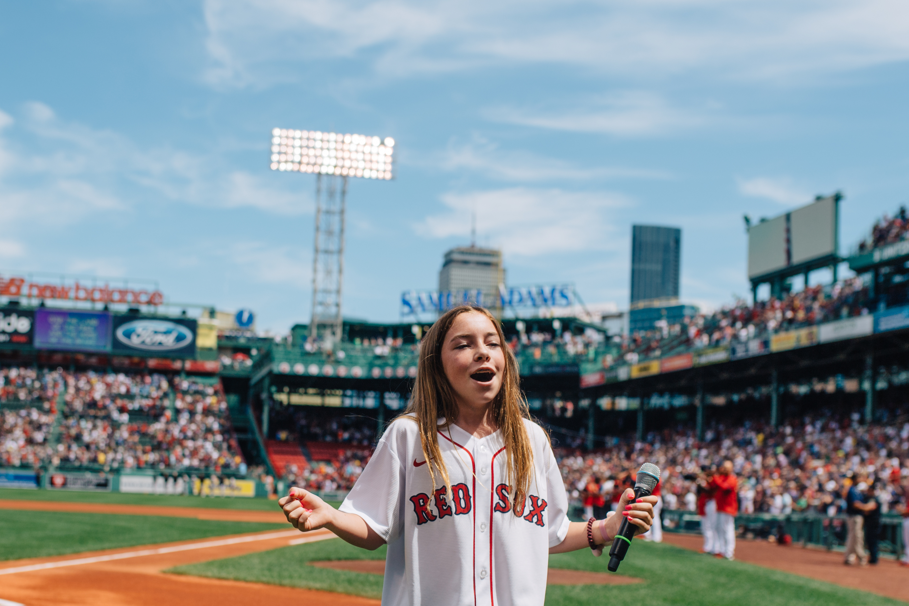 A girl wears a Red Sox jersey and holds a microphone, singing in Fenway Park.