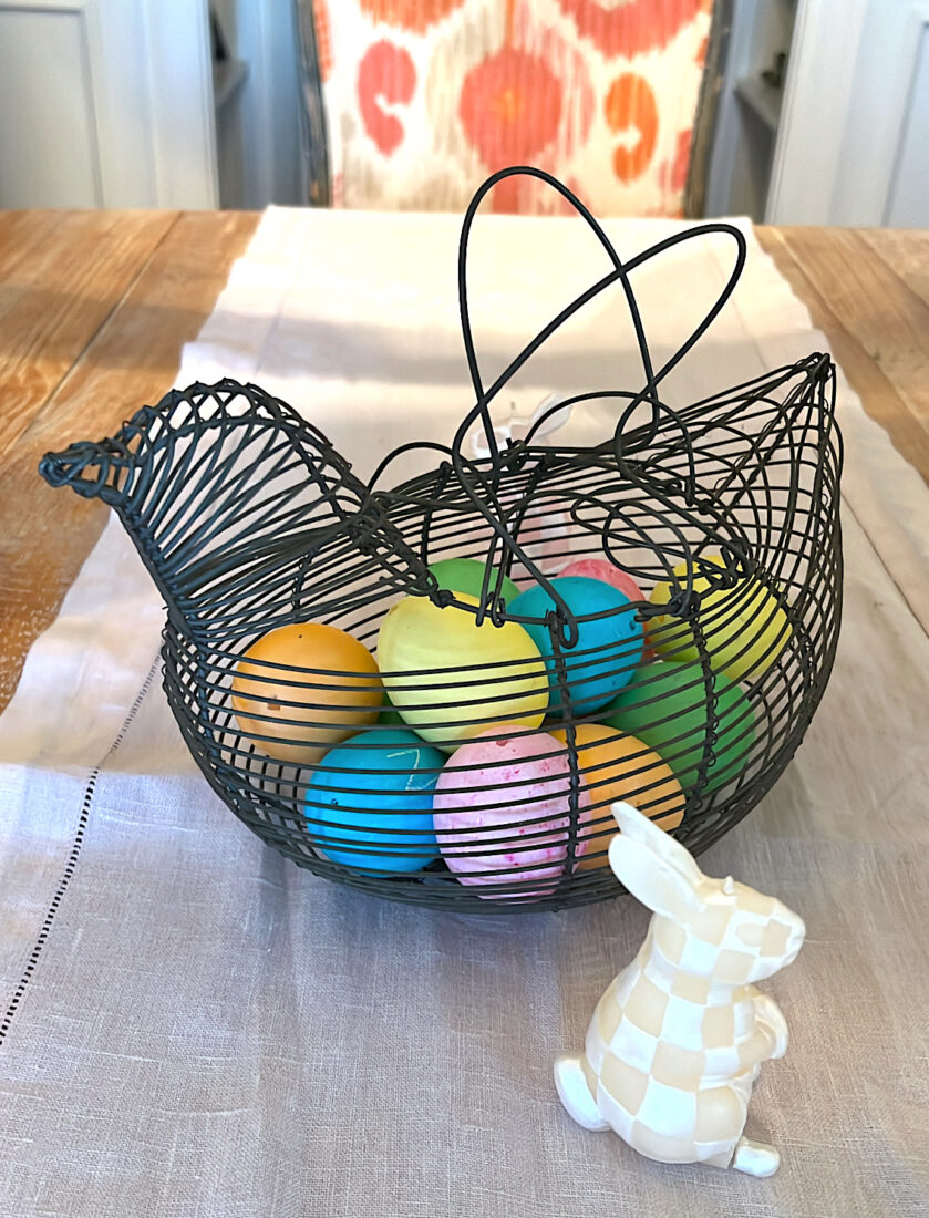 Colored easter eggs sit inside a metal chick-shaped basket. A small candle of a bunny sits to the right.