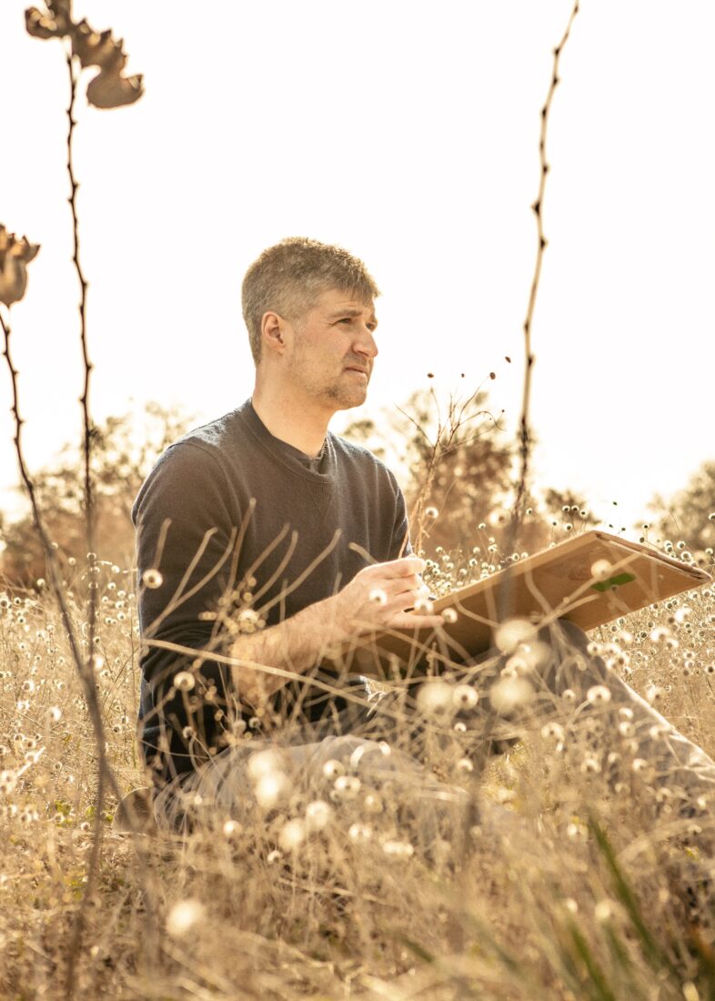 A man holds a board and paintbrush while sitting in a field