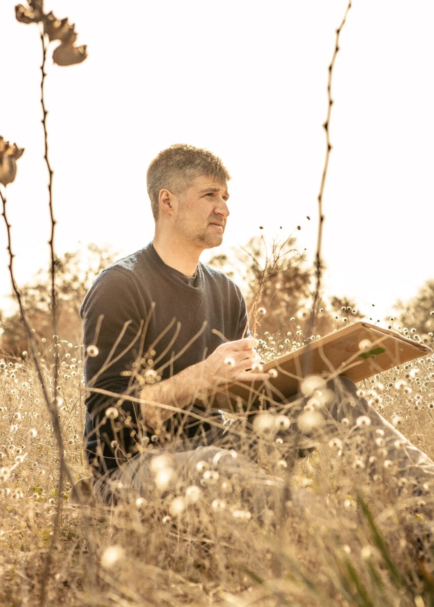 A man holds a board and paintbrush while sitting in a field