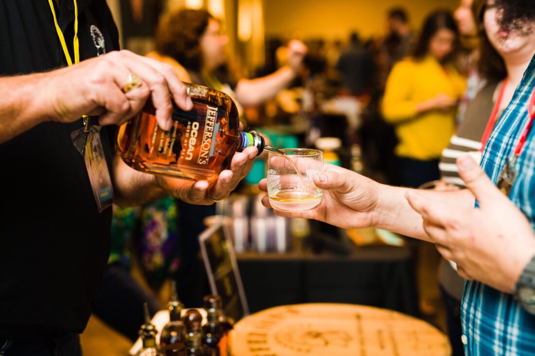 A bottle pours bourbon into a glass cup a person is holding.