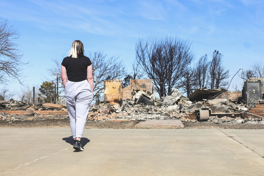 A girl wearing a black shirt and grey sweat pants stands in front of the ruins of a house burnt down from a wildfire.