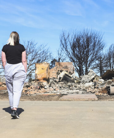 A girl wearing a black shirt and grey sweat pants stands in front of the ruins of a house burnt down from a wildfire.