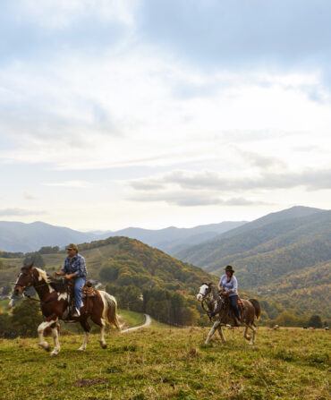 Two people on horseback ride over a ridge on a mountain with a grand landscape in the background
