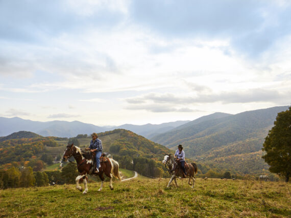 Two people on horseback ride over a ridge on a mountain with a grand landscape in the background