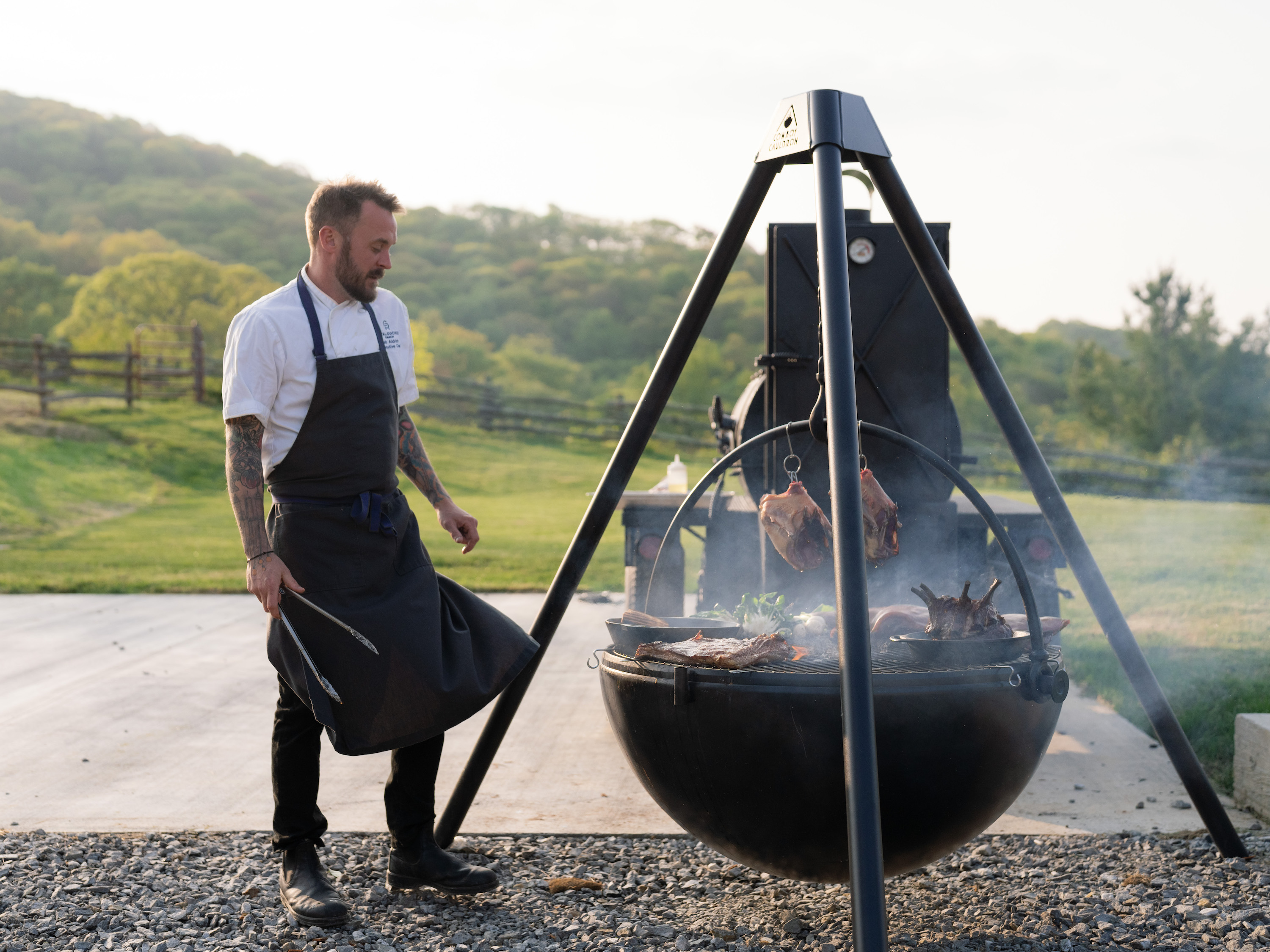 A chef stands by a large fire pit grill with food on it.