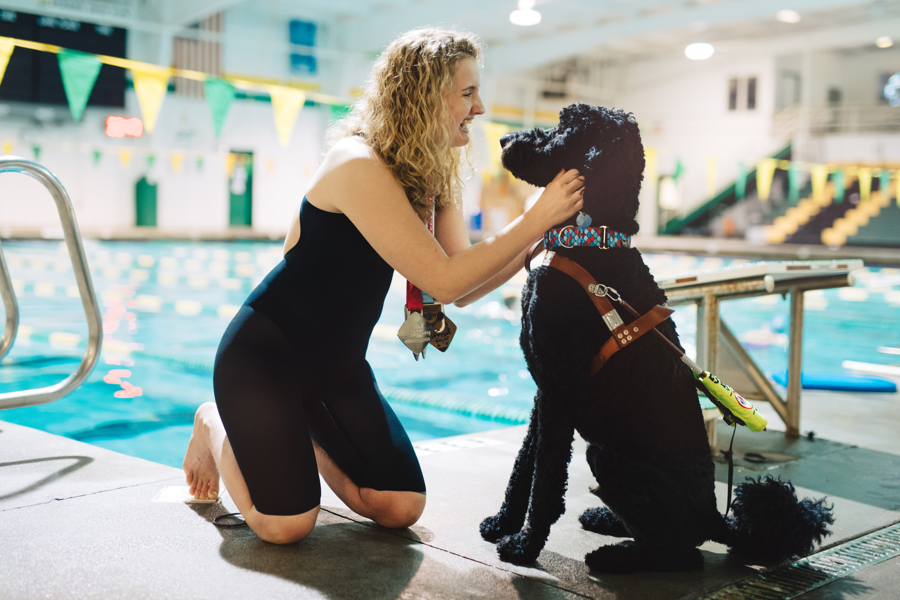 A young girl swimmer pets her service dog, a black poodle, at a pool.