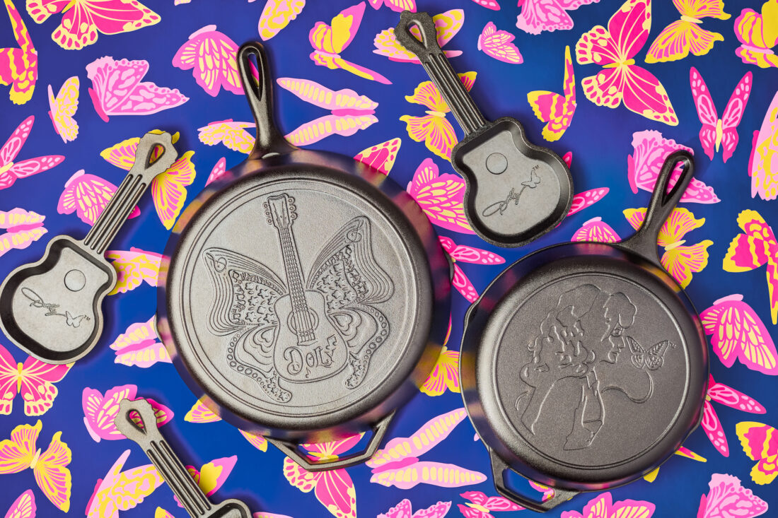 Four cast iron pans with etchings of butterflies, Dolly Parton's head and signature, and a guitar-shaped pan. They are on a blue, pink, and yellow butterfly-patterned background