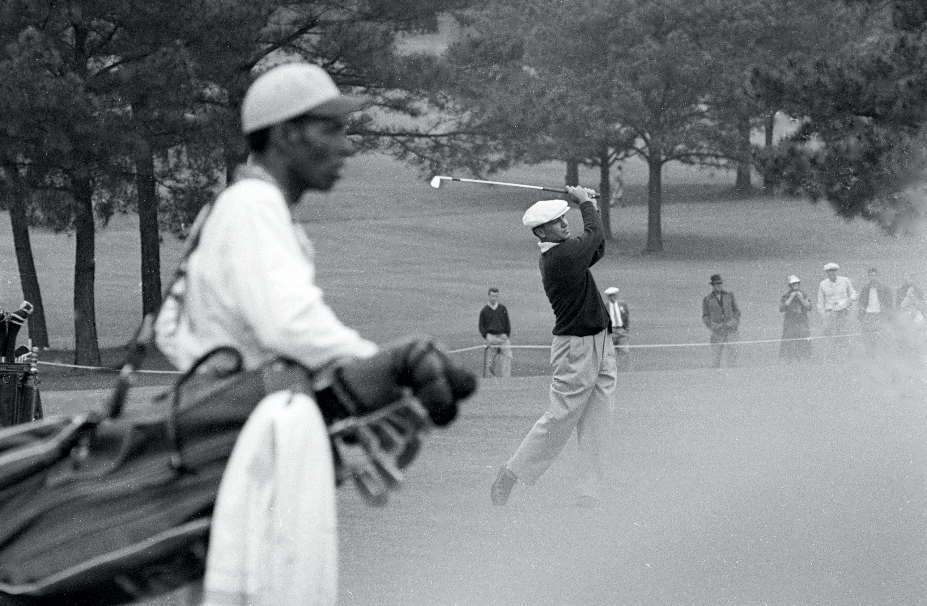 A Black caddie stands in the foreground as a man swings a golf club on the green