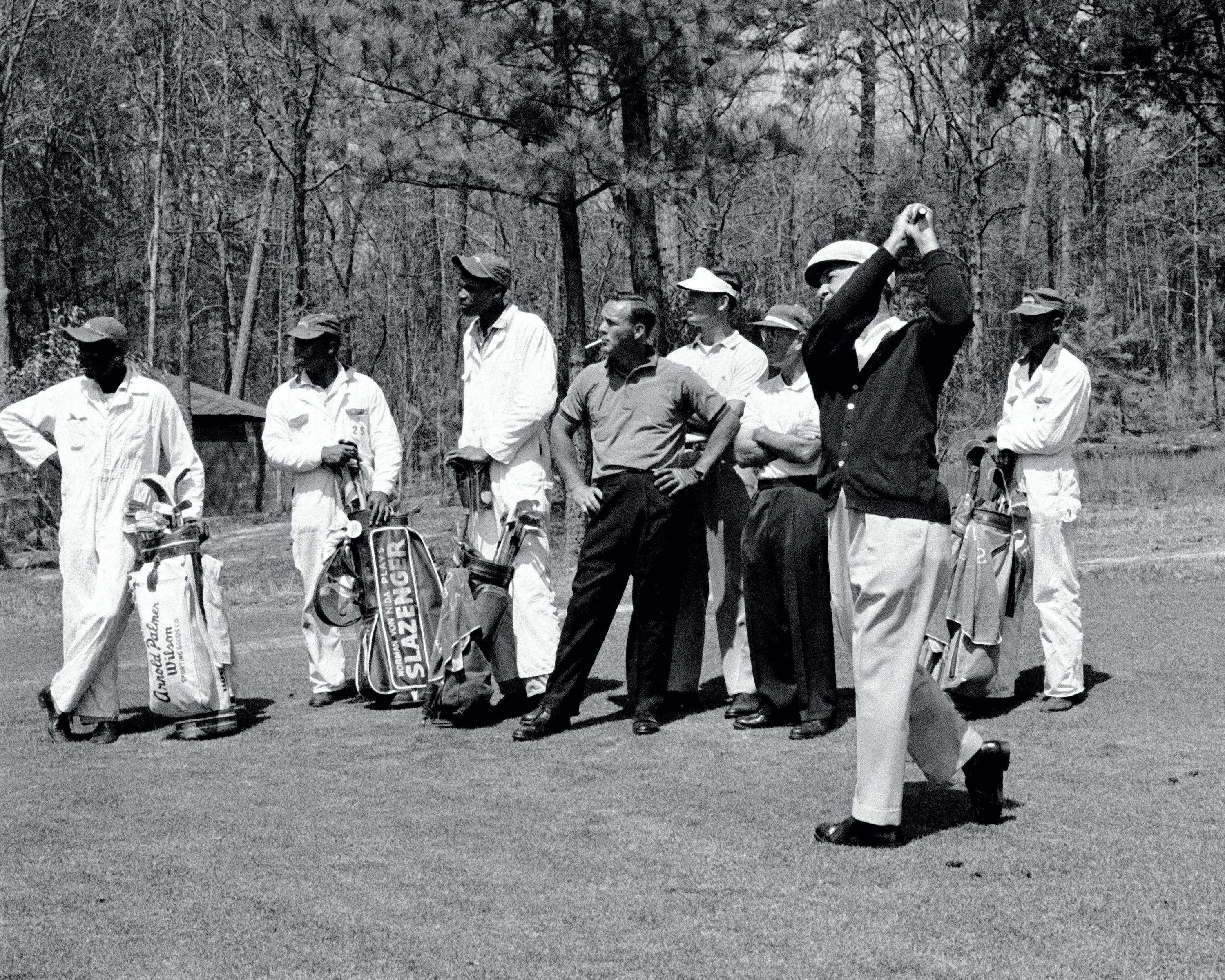 Arnold Palmer smokes and swings a club on the green with caddies around him.