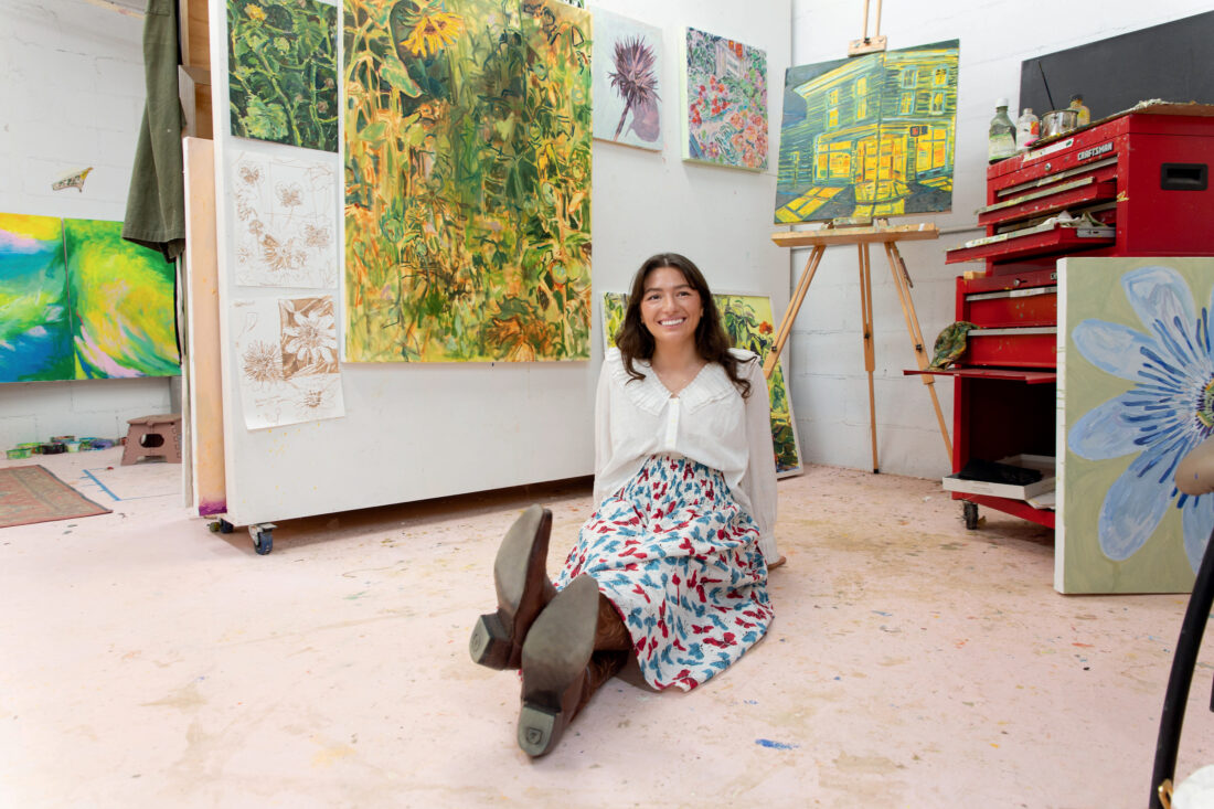 A girl sits on the floor in an art studio with paintings around her. She wears a blue, red, and white patterned skirt and a white blouse.