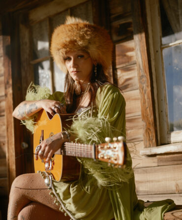A woman, wearing a fuzzy hat and a green feathery robe, holds a guitar on a wood porch