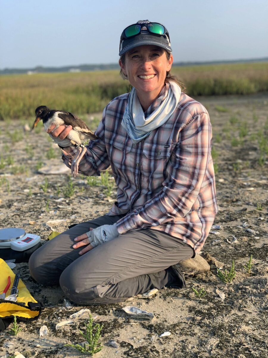 A woman holds a bird in her hand and kneels in the marshland
