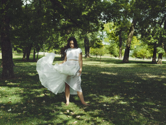 A woman twirling in a white flowy dress in a large wooded park.