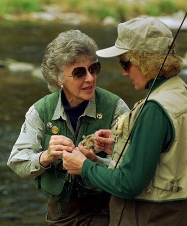 A woman teaches another woman how to attach a fly to a rod in a river