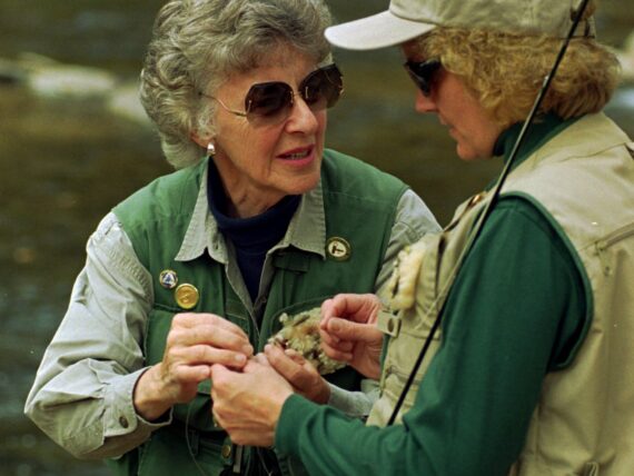 A woman teaches another woman how to attach a fly to a rod in a river