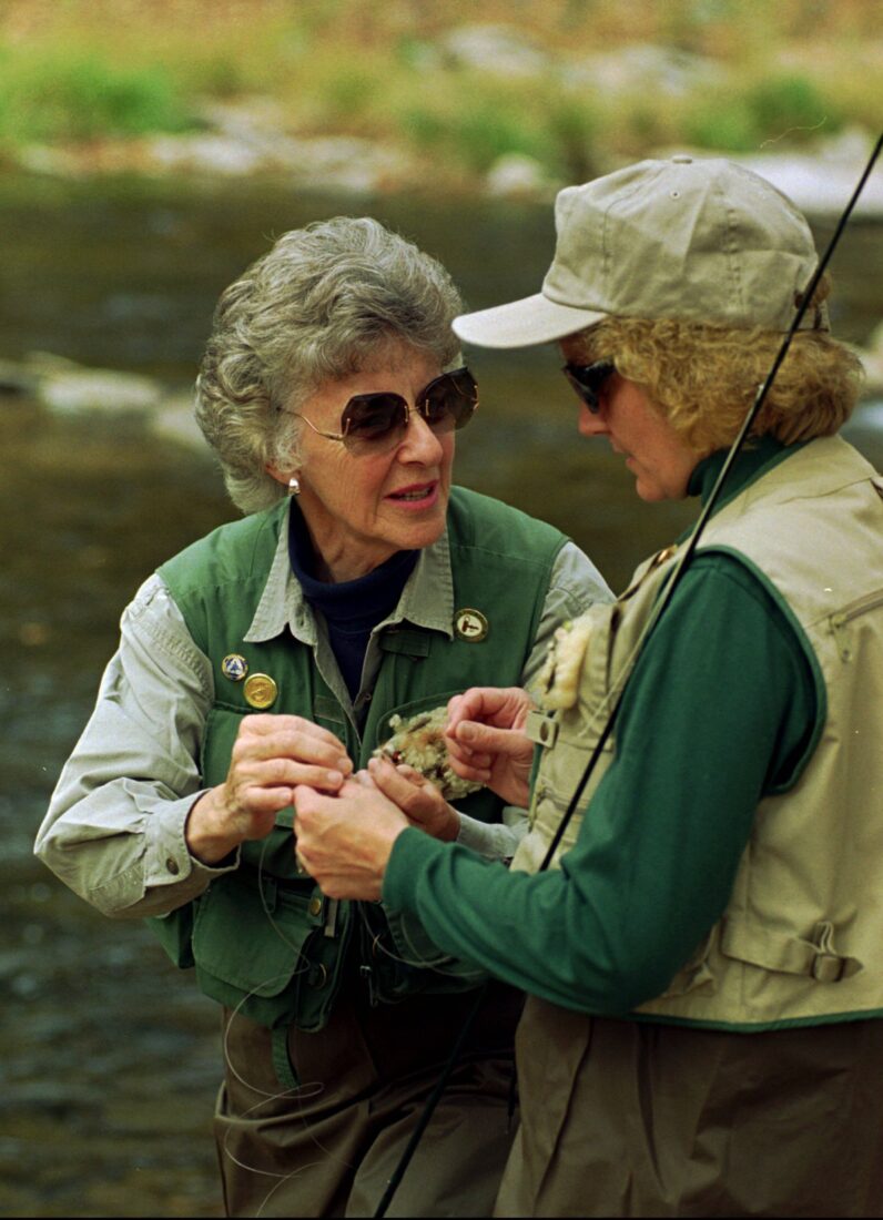 A woman teaches another woman how to attach a fly to a rod in a river